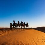 A group of travelers on camels crossing the sandy dunes of the Sahara Desert under a clear blue sky.