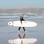 A surfer walks along Agadir beach carrying a surfboard, reflecting in wet sand.